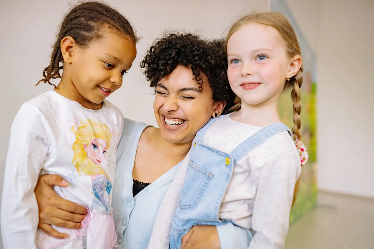 A smiling teacher embracing two young children in a bright classroom